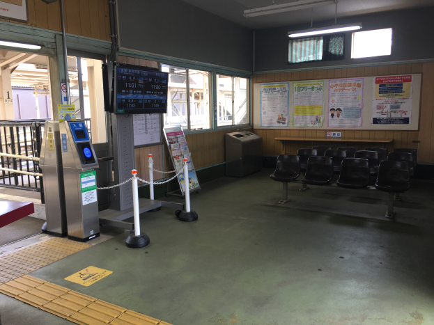 Ein belebter Bahnhofseingang mit Sitzgelegenheiten, einem Tresen, Ticketautomaten, Pfosten mit Ketten, Informationsanzeigen, Plakaten an den Wänden, Deckenleuchten und Geländern.