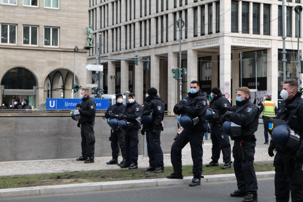 Eine Gruppe von Polizisten in schwarzen Uniformen und Masken steht vor einem Gebäude mit Glasfenstern und Säulen in Berlin, Deutschland, einige halten Helme, mit Laternenmasten, Verkehrsampeln, Schildern mit Text, einer Statue, anderen Menschen und einem grasbewachsenen Boden im Hintergrund.