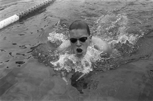 Ein Mann schwimmt in einem Pool mit offenem Mund und trägt eine Schwimmbrille, umgeben von Bahnmarkierungen, auf einem Schwarz-Weiß-Bild.