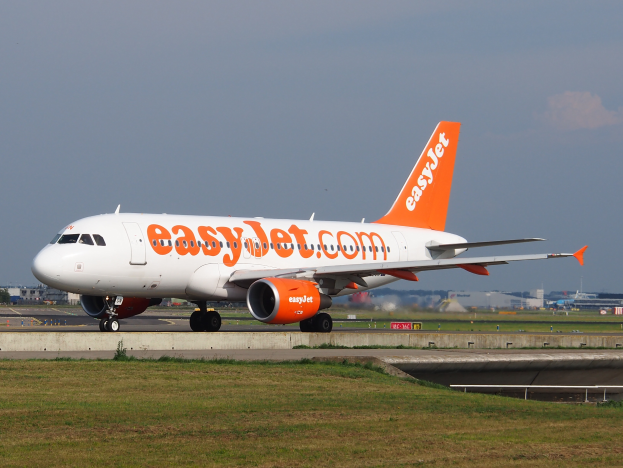Ein EasyJet Airbus A320-200 auf dem Rollfeld des London Heathrow Airport, umgeben von Gras und einer Straße im Vordergrund, sowie Gebäuden, Bäumen und einem klaren blauen Himmel im Hintergrund.