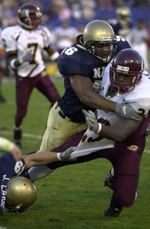 Zwei Fußballspieler in Helmen und Uniformen kämpfen auf dem Feld gegeneinander, während Teamkollegen und Zuschauer im Hintergrund zuschauen.
