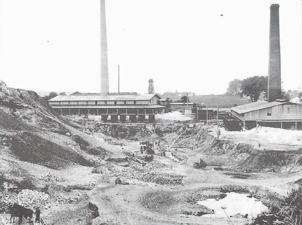 Ein Schwarz-Weiß-Foto einer Baustelle mit zwei Schornsteinen im Hintergrund, umgeben von Gebäuden, Masten, Bäumen und einem klaren Himmel, mit Text unten, der darauf hinweist, dass es ein Steinkohlekraftwerk ist.