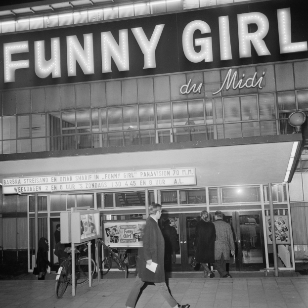 Eine Schwarz-Weiß-Szene von Menschen, die vor einem Gebäude mit einer Tafel 'Funny Girl du Midi' auf der Straße gehen, mit in der Nähe geparkten Fahrrädern.