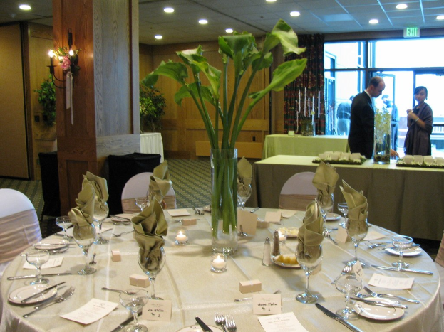 Ein Hotel-Essbereich mit einem weißgedeckten Tisch, auf dem Teller, Besteck, ein Glas und Zweige stehen, umgeben von Stühlen, einem grünen Tisch im Hintergrund, einem Mann in einem schwarzen Blazer, einem stehenden Mädchen, einer Tür mit der Aufschrift 'Ausgang' und zwei Männern dahinter.