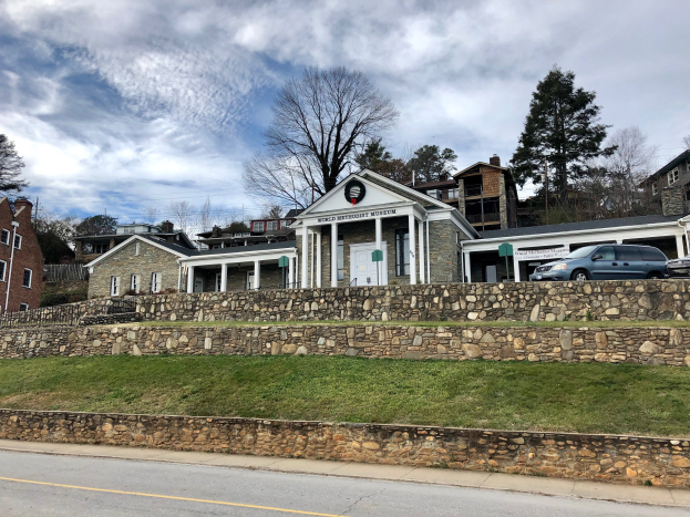 Steinwand mit parkenden Autos davor, flankiert von Gebäuden mit Fenstern und Türen, Bäumen und einem bewölkten Himmel, beschriftet als das kürzlich wiedereröffnete Weltkulturerbe-Museum.