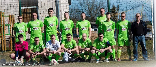 Eine Gruppe von Männern in grünen T-Shirts steht auf einem Fußballfeld, einige hocken, mit einem Ball vor ihnen in der Nähe eines Torpfahls; Gebäude, Bäume und Fahrzeuge sind im Hintergrund sichtbar.