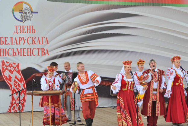 Eine Gruppe von Menschen in roter und weißer Kleidung performs auf der Bühne mit Mikrofonen und Instrumenten, mit einem "Ukrainischer Folklore-Festival"-Banner im Hintergrund.
