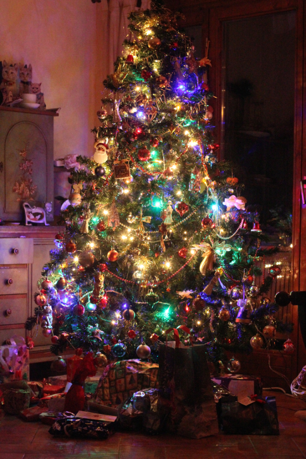 Ein Weihnachtsbaum mit Lichtern und Schmuck, Geschenke auf dem Boden, eine Kommode mit Gegenständen links und ein Fenster an der rechten Wand.