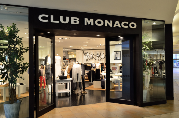 Außenansicht eines Club Monaco Geschäfts in einem Einkaufszentrum mit Glaswänden und -türen, Schaufensterpuppen in Kleidern, Auslagetischen mit Artikeln, einer Topfpflanze und Deckenleuchten.
