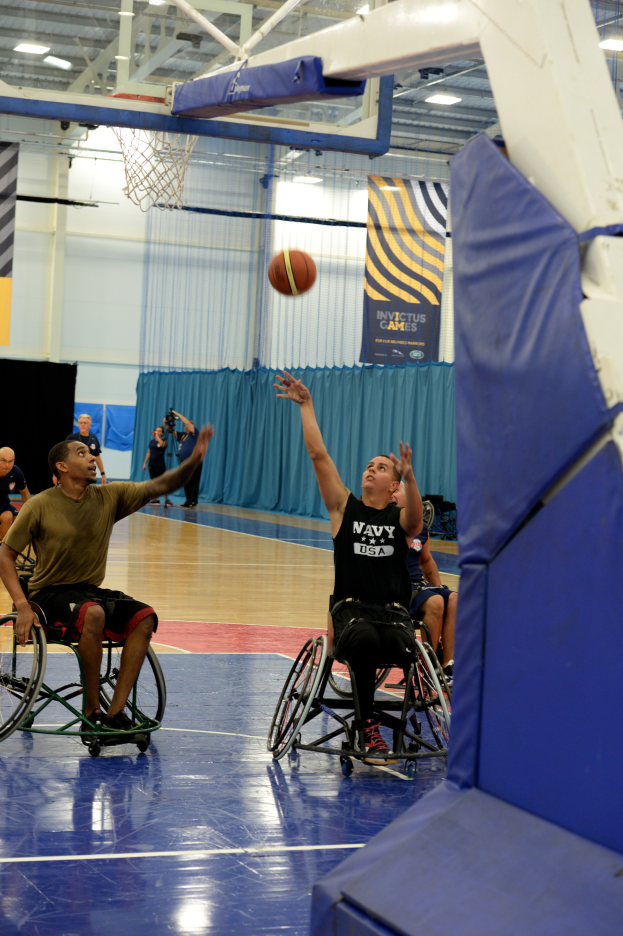 Eine Gruppe von Menschen in Rollstühlen, die Basketball in einer Turnhalle mit einem Basketballkorb, Bannern mit Text und Deckenleuchten spielen.