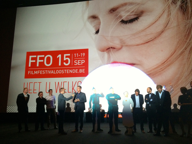 Eine Gruppe von Menschen auf einer Bühne mit einem Mann in der Mitte, der ein Mikrofon hält, ein Display hinter ihnen zeigt den Text "Foto Filmfestivaloostende 2015" und ein Bild einer Frau, sowie einige Menschen neben dem Display sitzen.
