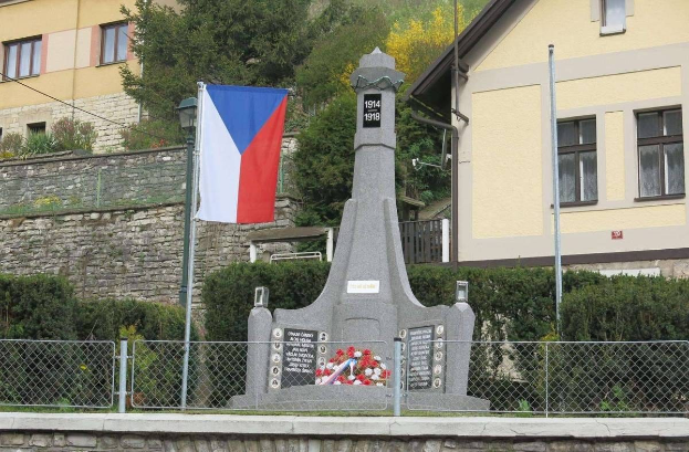 Ein Denkmal mit einer Fahne oben drauf, umgeben von einem Zaun, Pflanzen, Bäumen und einer Wand, vor einem Gebäude, als Mahnmal für die Opfer des Zweiten Weltkriegs, mit eingravierter Inschrift.