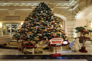 Ein große Weihnachtsbaum mit Schmuck, Lichtern und festlichen Deko-Elementen steht in der Lobby eines Hotels und ist umgeben von Geschenkboxen, einem Spielzeug, einer Tafel mit Text, Möbeln und einem Spiegel an der Wand.