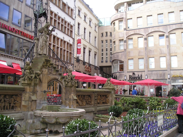 Eine Stadtansicht mit Gebäuden, Zelten und Menschen im Hintergrund und einer Skulptur, einem Teich, Pflanzen und Geländer im Vordergrund.