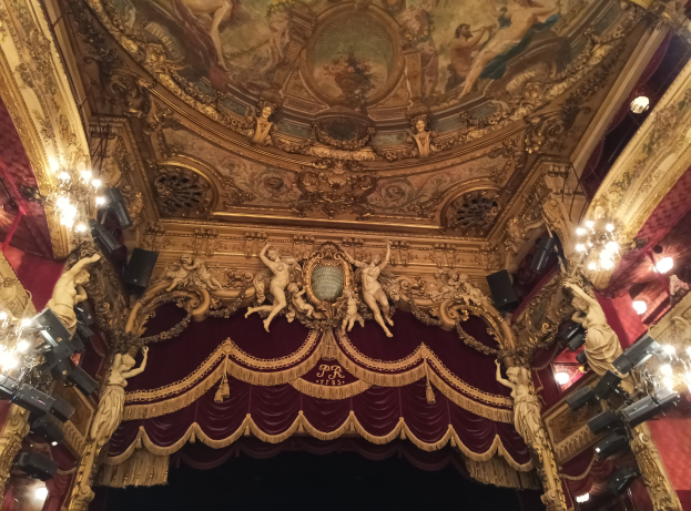 Theaterinnenraum mit roter Gardine, Kronleuchtern, Deckenplastiken, Lichtern und Gemälde, mit einer Bühne mit ein paar Leuten unten.