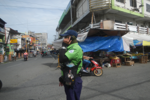Ein Polizeibeamter in grüner und blauer Uniform und blauer Mütze steht auf der Seite einer Straße und spricht auf einem Handy, mit Fahrzeugen, die auf der Straße fahren, Ständen mit verschiedenen Gegenständen auf seiner rechten Seite und einem bewölkten Himmel mit einem Gebäude mit Fenstern im Hintergrund.
