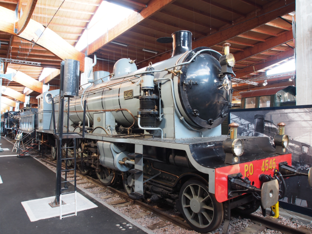 Alte Dampflokomotive in einem Museum ausgestellt, umgeben von Menschen auf einem Bahngleis unter metallenen Deckenleuchten, mit einer Informationstafel auf der rechten Seite.