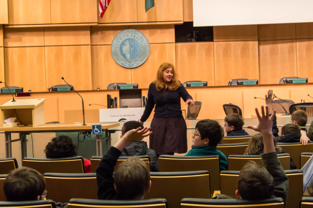 Eine Frau, die als Stadtoberhaupt von Seattle identifiziert wird, steht an einem Podium mit Mikrofonen in einem Klassenzimmer voller aufmerksamer Kinder, mit einem Logo und zwei Flaggen an der Wand dahinter.