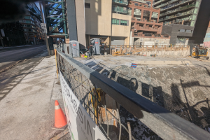 Eine Baustelle mit einem Zaun und einem Verkehrskegel im Vordergrund, eine Straße links, Gebäude im Hintergrund, ein Baumaschinenfahrzeug und ein Müllcontainer.