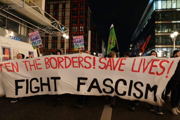 Eine Gruppe von Menschen marschiert mit einem Transparent, auf dem "Ten the Borders Save Lives, Fight Fascism" steht, durch die Straße, mit Laternenmasten, Gebäuden und einem sichtbaren Himmel im Hintergrund.