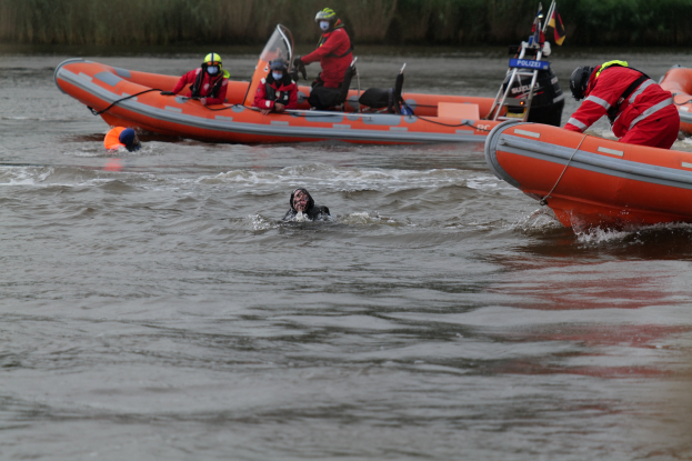 Gruppe von Menschen in einem aufblasbaren Boot auf einem Fluss, mit zwei Individuen im Wasser im Vordergrund und Vegetation im Hintergrund, alle tragen Schwimmwesten und Helme während einer Rettungsoperation.