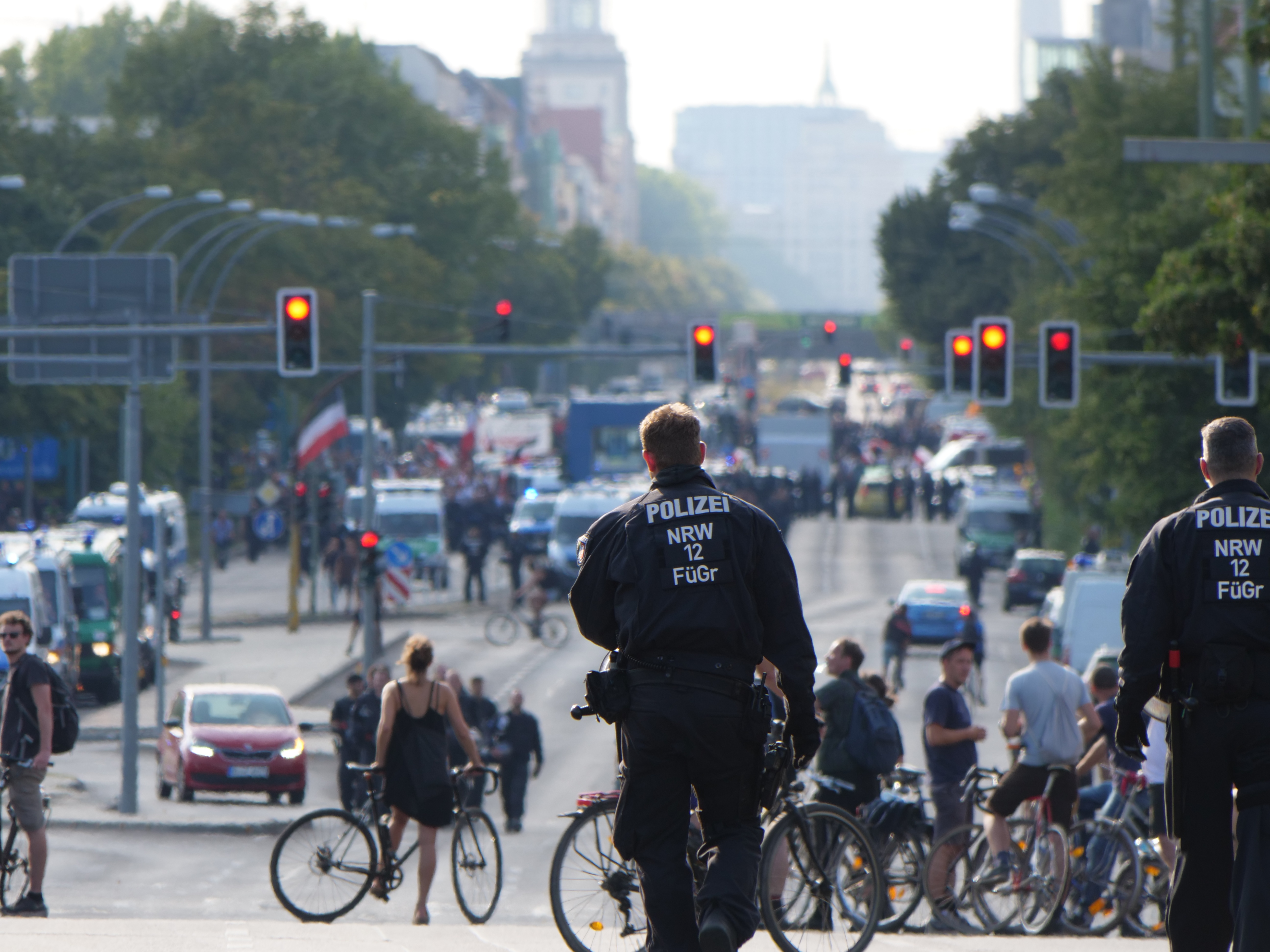 Zwei Polizeibeamte stehen an einer Straße neben einer Gruppe von Radfahrern, mit Fahrzeugen, Verkehrszeichen, Bäumen, Gebäuden und einem klaren blauen Himmel im Hintergrund.