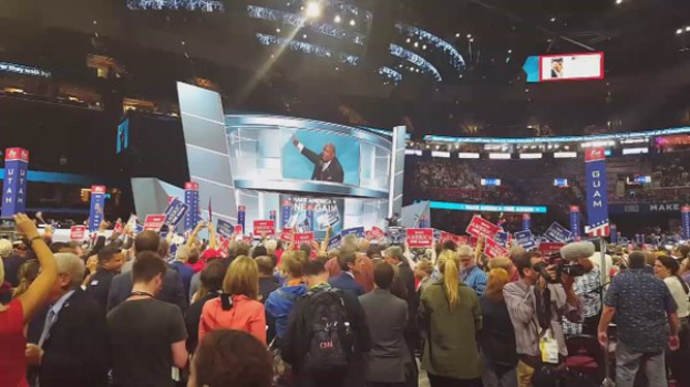 Eine große Menschenmenge steht vor einer Bühne, hält Schilder und Transparente mit Text, während eine Person eine Kamera in der foreground hält und Bildschirme und Scheinwerfer im Hintergrund zu sehen sind bei der Republican National Convention.