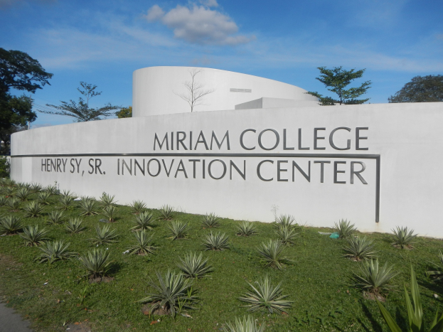 Außenansicht des Miriam College Innovation Center mit einer textgeschmückten Wand, umgeben von Grün, einem Weg, Bäumen und einem bewölkten Himmel.