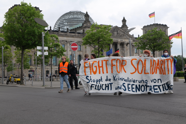 Eine Gruppe von Menschen marschiert auf einer von Bäumen gesäumten Straße und hält ein Banner mit der Aufschrift 'Kampf für Solidarität' hoch, mit parkenden Fahrzeugen und einem mehrstöckigen Gebäude im Hintergrund unter einem bewölkten Himmel.