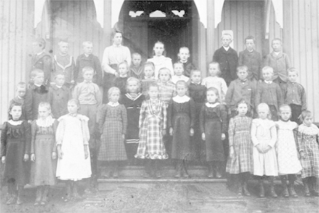 Ein Schwarz-Weiß-Foto einer Gruppe von Kindern, die vor einem Gebäude mit einer Tür und Säulen stehen, mit dem Text "Erste Klasse" unten.