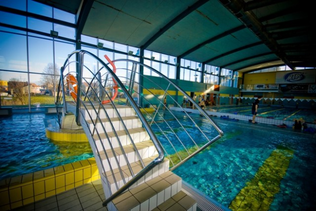 Große Hallenbad mit Treppe, die hineinführt, Menschen im Wasser, Wand mit Text auf der rechten Seite, Metallstäbe an der Decke und Hintergrund mit Bäumen, Gebäuden und einem klaren blauen Himmel.