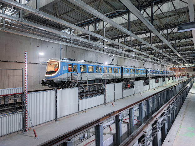 Ein blauer und weißer Zug fährt durch ein großes Gebäude mit einer Plattform auf der rechten Seite und einer Decke mit Stangen und Lampen darüber, Teil eines neuen Schienensystems in Deutschland.