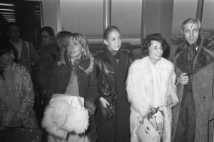 Eine Gruppe von Menschen in Jacken, einige halten Taschen, steht zusammen in einem Flughafen mit einer Wand im Hintergrund, dargestellt in Schwarz-Weiß.