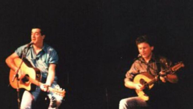 Zwei Personen sitzen auf einem Stuhl, eine spielt Gitarre und die andere singt in ein Mikrofon.