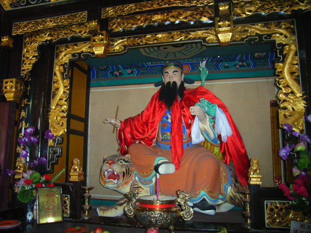 Eine Statue eines chinesischen Mönchs, der bunt gekleidet auf einem Tiger sitzt, in einem Bogen mit goldenen Verzierungen, vor ihm eine Vase und in der Mitte eine Schüssel.
