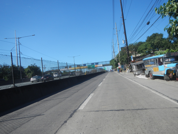 Arterielle Straße mit fahrenden Fahrzeugen, geparktem Bus, Strommasten mit Drähten, einem Zaun, Bäumen, Laternen, einer Brücke im Hintergrund und einem blauen Himmel.