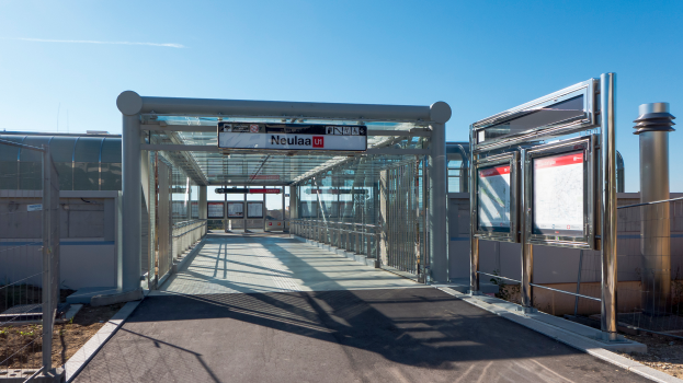 Ein Gehweg mit Geländern und Pfosten führt zu einem Bushaltestellenschild, mit Texttafeln an den Seiten und Gebäuden unter einem klaren blauen Himmel im Hintergrund.