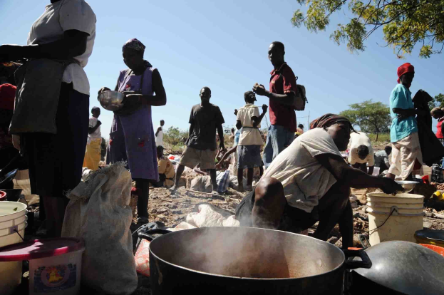 Eine Gruppe von Menschen bereitet in einer ländlichen Gegend unter freiem Himmel Essen zu, mit einem großen Topf, Eimern und Abdeckungen auf dem Boden, unter einem klaren blauen Himmel.