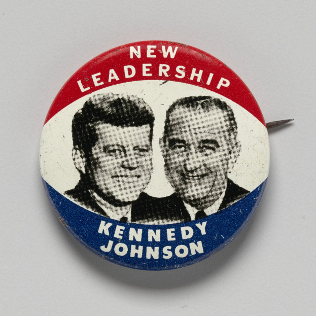 Ein Ansteckbutton auf einer weißen Oberfläche zeigt zwei Bilder von Kennedy und Johnson mit den Wörtern "Neue Führung" darunter.