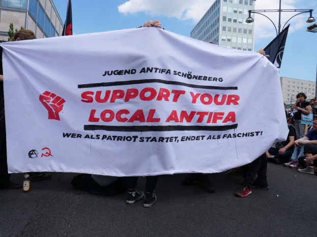 Eine Gruppe von Menschen mit einem Banner, auf dem "Support Your Local Antifa" steht, und einer Flasche auf einer Straße, mit Gebäuden, Straßenlaternen, Lichtern, Bäumen und einem bewölkten Himmel im Hintergrund.