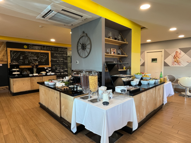 Ein Restaurant-Innenraum mit Tischen und Stühlen, Speisen, einer Tafel mit Text, einer Wanduhr, Deckenleuchten, einer Klimaanlage und einer Tür im Hintergrund, Located in the Hotel de Ville in Paris.