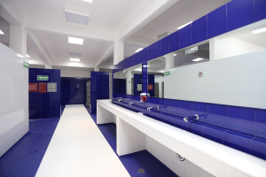 Öffentliche Toilette mit blauen Kacheln, weißen Arbeitsplatten, Waschbecken mit Wasserhähnen, einer Plattform mit einer Flasche und anderen Gegenständen, Texttafeln an den Wänden, Deckenleuchten und Säulen.