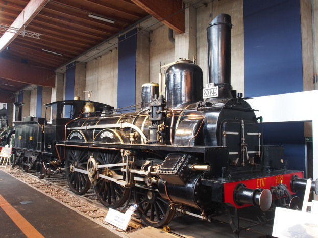 Alte Dampflokomotive in einem Museums Setting mit umgebenden Eisenbahnschienen, Informationsschildern und anderen Ausstellungen an einer Wand mit Deckenbeleuchtung.