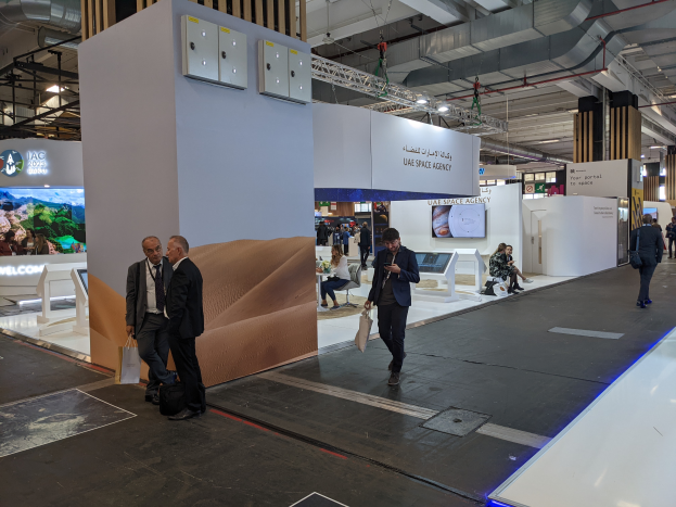 Eine Gruppe von Menschen geht um den Messestand der UAE Space Agency herum, mit Taschen tragend, Informationsplakate, Bildschirme und andere Gegenstände im Hintergrund, unter Deckenleuchten und Säulen.