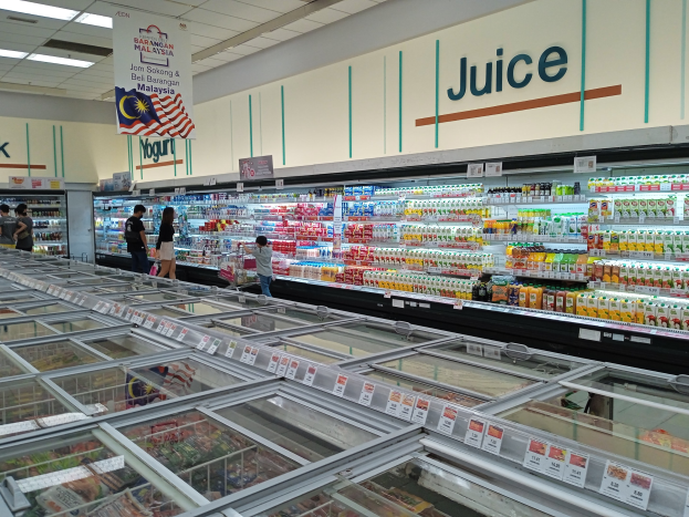 Ein hell erleuchteter Saftladen in einem Supermarkt zeigt eine Auswahl an Flaschengetränken, frischem Gemüse und verpackten Waren auf Regalen, mit einigen Einkäufern und einer Deckenbeleuchtung, die den Raum erhellt.