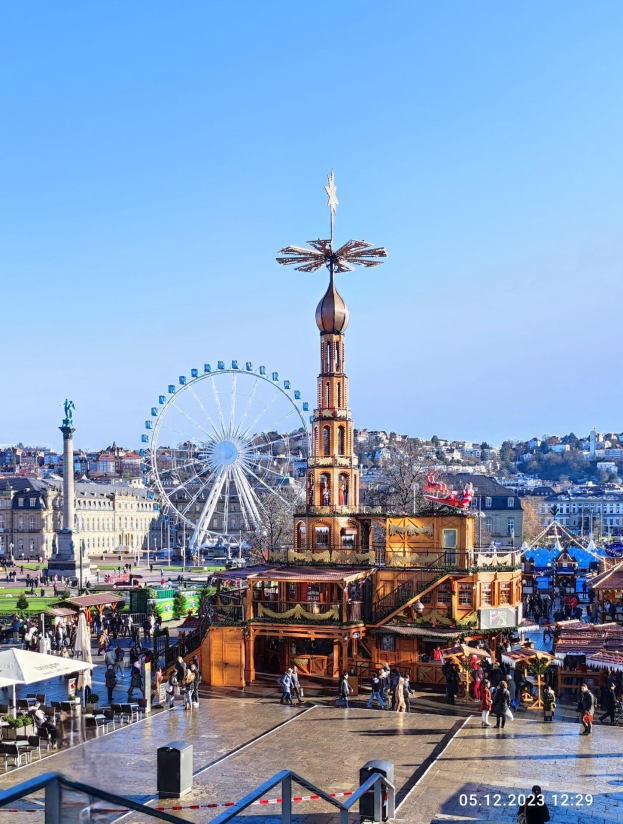 Ein belebter Stadtplatz mit einem zentralen Riesenrad, umgeben von Gebäuden, Bäumen und Menschen unter einem klaren blauen Himmel, mit Marktzelten und Geländern, mit einem Wasserzeichen unten.