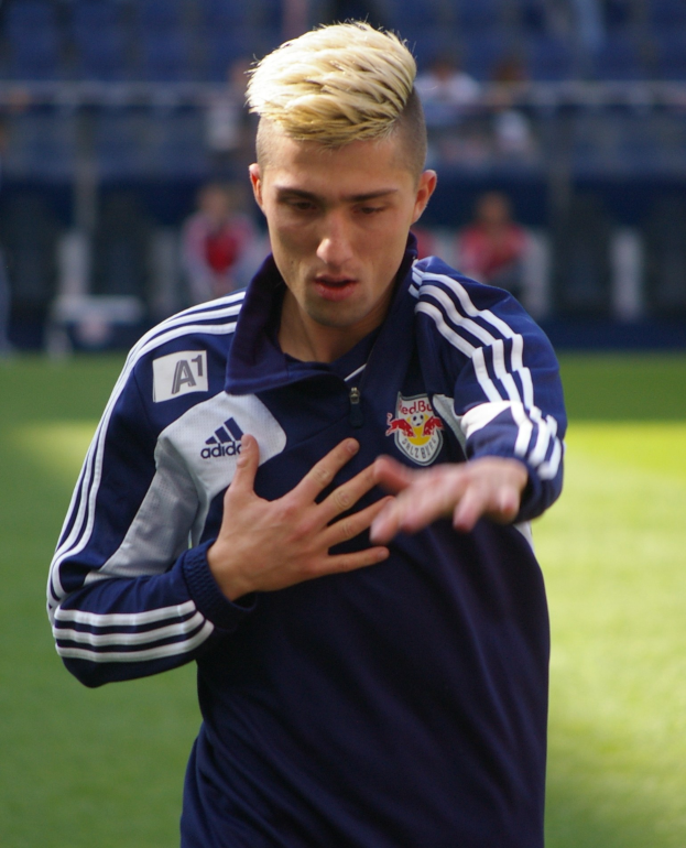 Ein junger Mann in einem blauen T-Shirt steht auf einem Fußballfeld und hat die Arme ausgestreckt und einen entschlossenen Gesichtsausdruck, unscharfer Hintergrund.