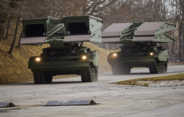Zwei Militärfahrzeuge fahren nebeneinander auf einer Straße mit einer Person in einem der Fahrzeuge, umgeben von Gras, Bäumen und einem klaren blauen Himmel.