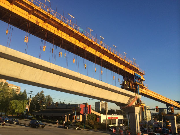 Eine Baustelle in der Mitte mit Fahrzeugen und Gebäuden links, unter einem blauen Himmel.