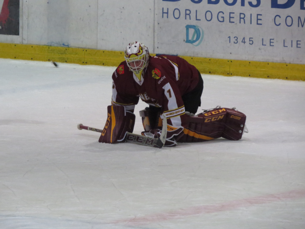 Eishockey-Spieler in rot-gelber Uniform, der einen Schuss auf dem Eis hält, vor einer Wand mit Text im Hintergrund.